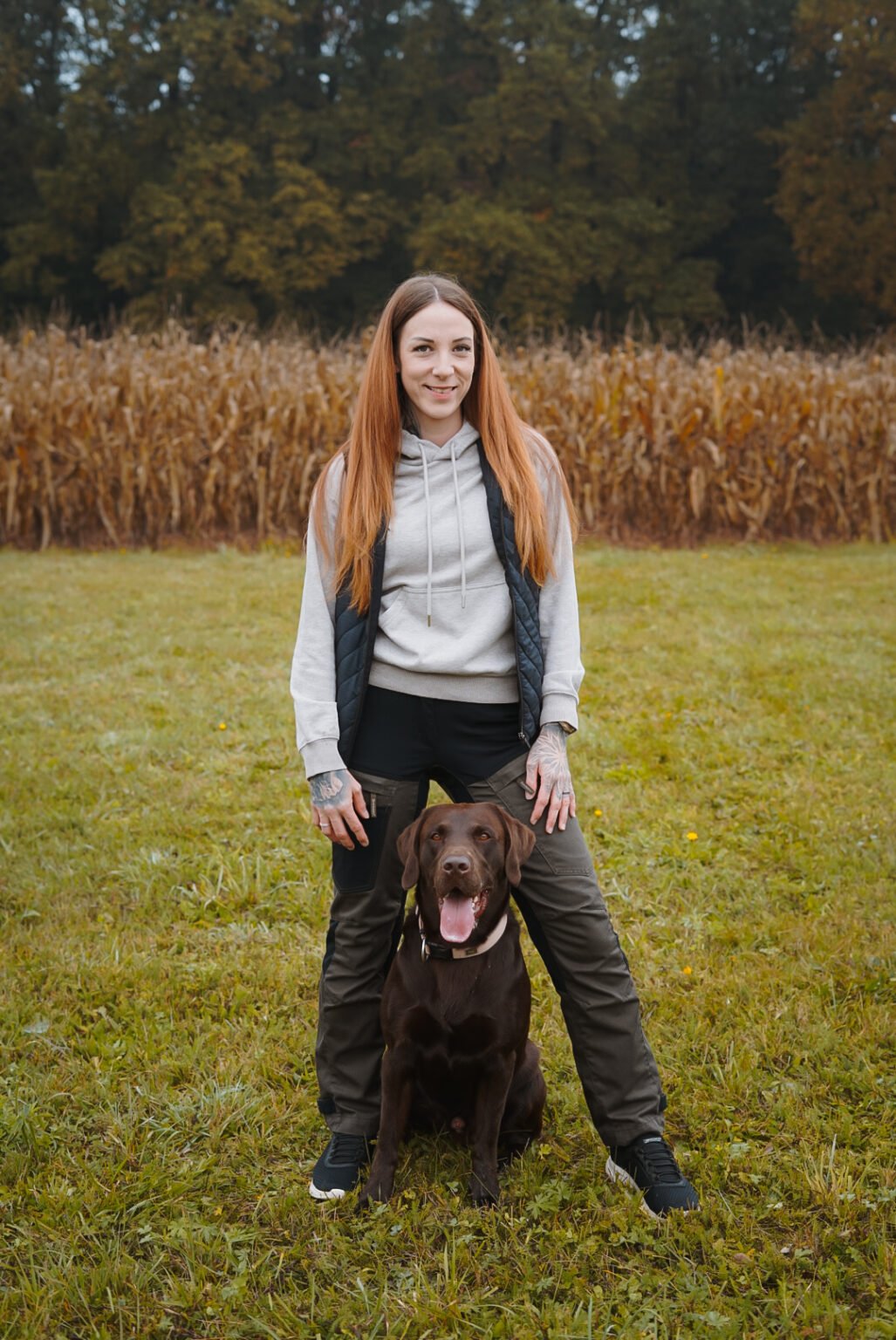 Hundetrainerin Natascha Kelz (Tascha) steht lächelnd auf einer Wiese vor einem Maisfeld, ihr brauner Labrador Aaron sitzt zwischen ihren Beinen