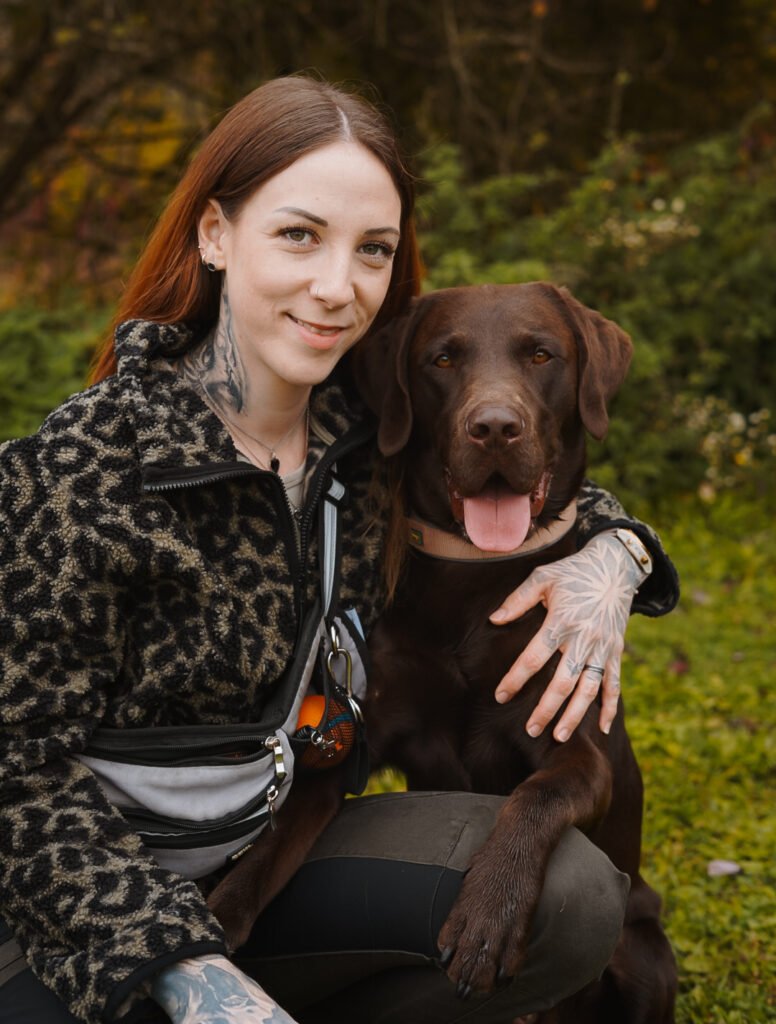 Hundetrainerin Natascha Kelz (Tascha) kniet lächelnd neben ihrem braunen Labrador Aaron und hat den Arm um ihn gelegt